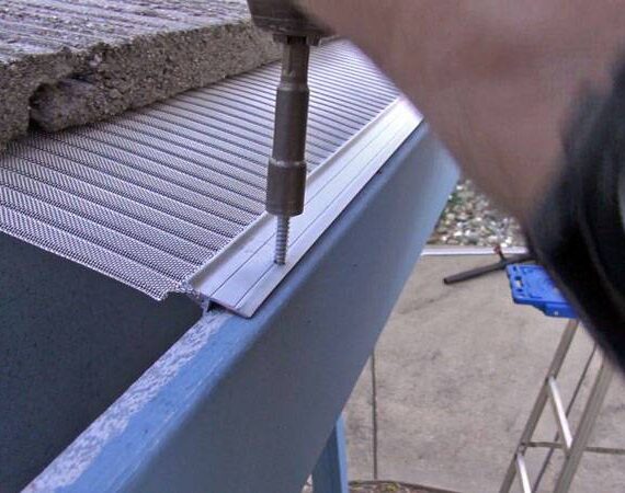 A person installs a stainless steel micro-mesh gutter guard with V-Bend Technology on a roof edge using a power drill. Part of the roof and mesh guard are visible, helping to protect against pine needles, with a concrete slab above.